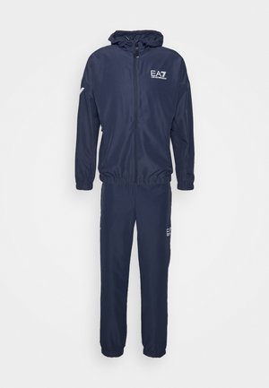 Trening de culoare bleumarin, realizat dintr-un material ușor, care include o jachetă cu fermoar, glugă și manșete elastice, asociată cu pantaloni tapered asortați.