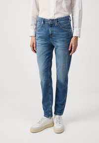 Niebieskie dżinsy o slim fit, z pięcioma kieszeniami, delikatnym wyblaknięciem i kontrastowym przeszyciem, w połączeniu z białymi sneakersami i jasną koszulą.