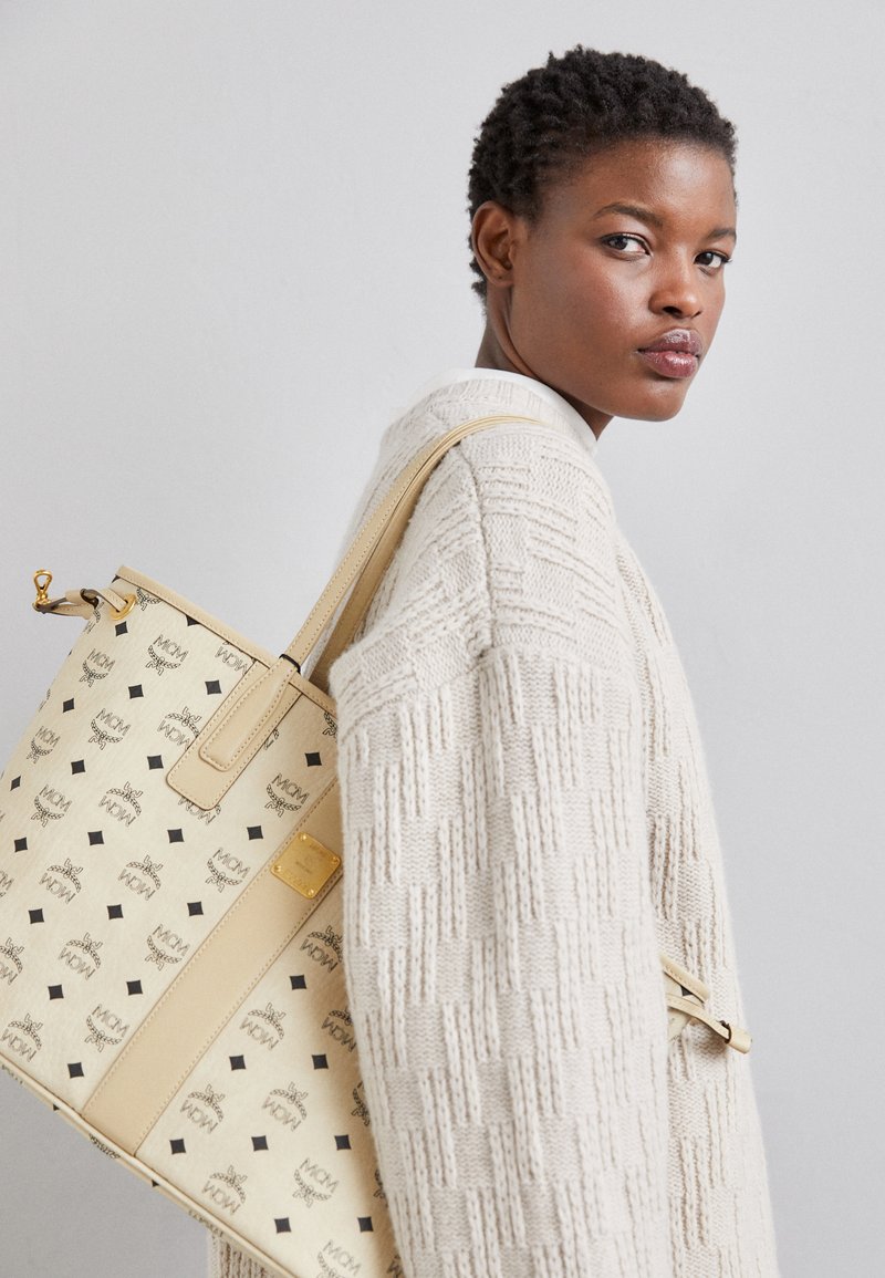 Sac fourre-tout en cuir beige, arbore un motif avec des logos noirs et des accents en forme de diamant, complété par un détail en métal doré.