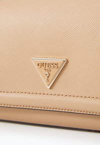 Bežová textura kabelka s pozlacenou trojúhelníkovou logoplatem s nápisem "GUESS" a otazníkem. Hladké švy podél okrajů.