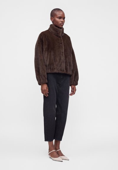 Femme portant une veste en fausse fourrure marron foncé, un pantalon court noir et des escarpins beige clair à bout pointu et talon bas, debout devant un fond clair uni.