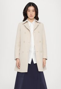 Beige dobbeltbrystet frakke lavet af stof, med krave, forreste knapper og sidelommer, båret over en hvid skjorte og en marineblå nederdel.