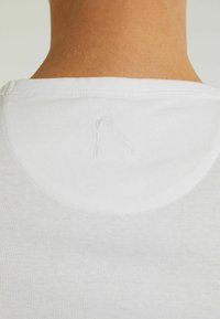 Bílé bavlněné tričko s kulatým výstřihem. Na zadní části krku má jemnou výšivku, která ukazuje minimalistický design.