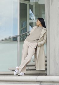 Beige trenchcoat, beige sweater og brede bukser. Hvide sneakers med farverige accenter. Står på trin nær glasdøre.