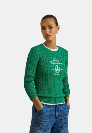 Donna con capelli scuri raccolti indossa un maglione verde lavorato a trecce con scritta logo bianca e jeans blu, mani in tasca, su sfondo semplice.