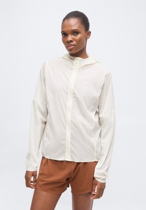 Frau trägt eine hellbeige Kapuzenjacke mit Reißverschluss und braune Shorts, steht mit neutralem Gesichtsausdruck vor einem einfachen Hintergrund.