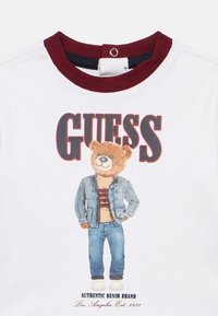 Λευκό βαμβακερό μπλουζάκι με γραφικό ενός αρκούδου που φοράει τζιν μπουφάν πάνω από μπεζ πουκάμισο και μπλε τζιν. Λεπτομέρεια με μπορντό ριμπ στην ψηλή λαιμόκοψη.