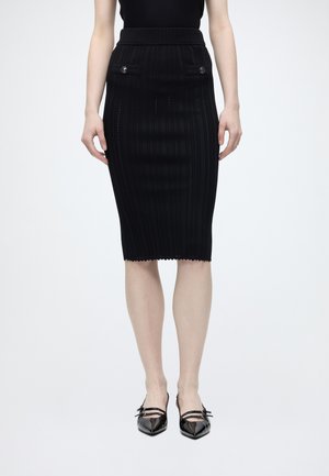 Jupe crayon noire longueur genou avec motifs verticaux subtils, deux rabats de poche avant avec boutons, portée avec des escarpins noirs à bout pointu avec brides.