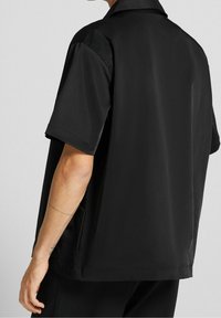 Czarna koszula z krótkim rękawem, wykonana z gładkiej tkaniny o luźnym kroju. Posiada kołnierzyk z klapą oraz boczne rozcięcia dla swobody ruchów.