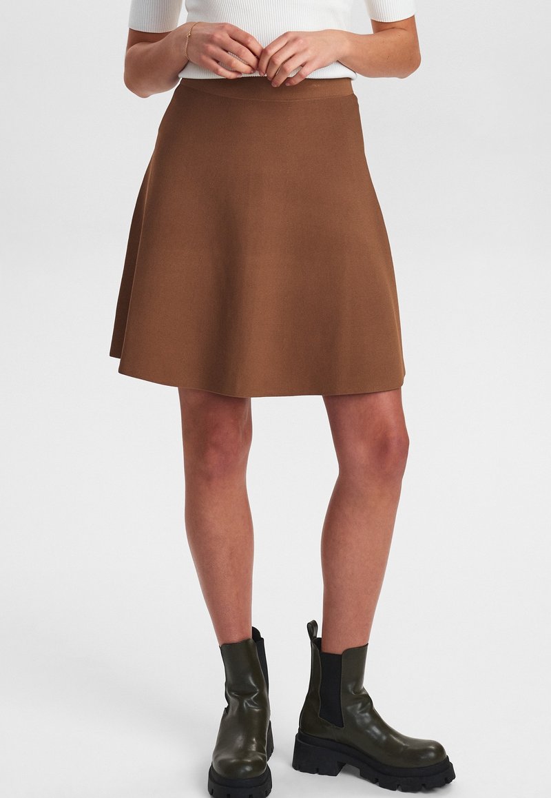 Femme portant une jupe marron jusqu'aux genoux, un haut blanc à manches courtes et des bottines noires épaisses, debout sur un fond uni.