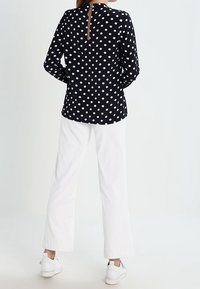Blusa nera a pois con maniche lunghe e zip sul retro, abbinata a pantaloni bianchi a palazzo e sneakers bianche. Texture del tessuto liscia.