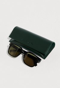 Črna pravokotna sončna očala s temnimi lečami poleg temno zelene usnjene etuije Lacoste na belem površju.