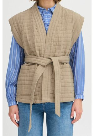 Person iført en beige quiltet ærmeløs vest med bælte over en blåstribet skjorte med knapper og blå jeans.