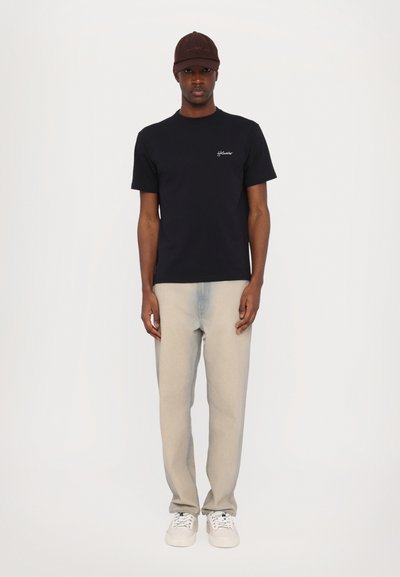 Black short-sleeve t-shirt with white logo, paired with light gray jeans and white sneakers. Brown cap completes the outfit.