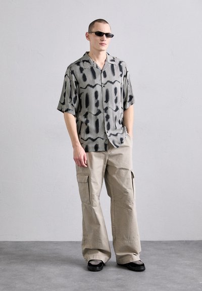 Chemise à manches courtes à rayures noir et blanc avec des formes abstraites. Pantalon cargo beige loose avec des poches latérales. Lunettes de soleil noires.
