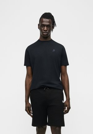 Homme portant un t-shirt noir à manches courtes avec un petit logo sur la poitrine et un short noir à cordon, debout devant un fond clair uni.