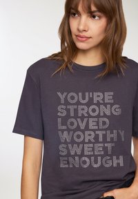 T-shirt grigio scuro a maniche corte con testo in rilievo argentato che dice "SEI FORTE, AMATO, MERITEVOLE, ABBASTANZA DOLCE." Materiale in cotone morbido.