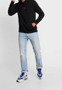 Sweat à capuche noir avec une poche kangourou, détail logo rouge, associé à un jean bleu clair déchiré et des baskets bleu et blanc.