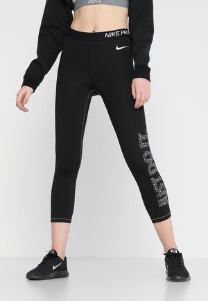 Kobieta nosząca czarne legginsy Nike Pro z białym logo na pasku oraz nakrapianym napisem „Just Do It”, zestawione z czarnymi sneakersami Nike.