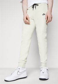 Ljusgrå joggingbyxor med svart dragsko, åtsittande mudd, sidofickor och en liten svart Nike-logotyp på vänster lår.