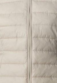 Světlý béžový puffer vest s horizontálním švem, předním zipovým uzávěrem a hladkým, lesklým povrchem. Obsahuje postranní kapsy.