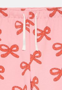 Rosa Jogginghose mit einem sich wiederholenden Design von glitzernden roten Schleifen. Verfügt über einen Kordelzugbund und weichen, strukturierten Stoff.