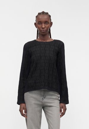 Kvinde med flettet hår iført tekstureret sort top med lange ærmer og lysegrå jeans, står mod en ensfarvet lys baggrund.