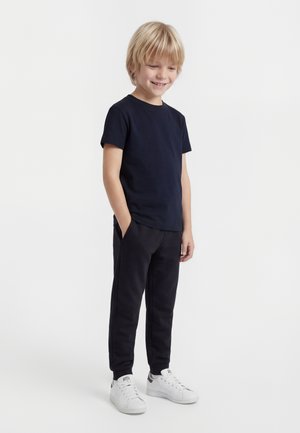 Ragazzo biondo in piedi con le mani in tasca, indossa una maglietta blu scuro, pantaloni neri e sneakers bianche su uno sfondo semplice.