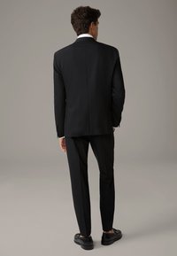 Giacca di un elegante completo nero con uno spacco singolo, abbinata a pantaloni slim-fit. Il colletto di una camicia bianca spunta da sotto la giacca. Scarpe eleganti nere.