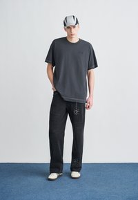 Junger Mann, der ein schwarzes übergroßes T-Shirt, schwarze zerrissene Jeans mit Nieten-Designs, weiße Turnschuhe und eine schwarz-weiße Kappe trägt, steht vor einem einfarbigen Hintergrund.