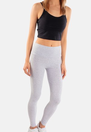 Kobieta w czarnym krótkim topie, jasnoszarych legginsach i białych trampkach stoi na tle jednolitego białego tła.