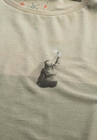 T-shirt verde oliva in cotone, con una silhouette nera di una figura con un pennello sul davanti, dalla texture morbida.