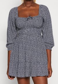 Námořnická modrá a bílá květinová šaty s čtvercovým výstřihem, nabíranými rukávy a přiléhavým pasem. Vyrobené z lehkého materiálu s volánkovou sukní.