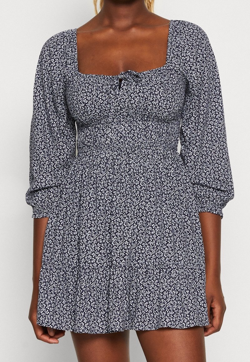 Námořnická modrá a bílá květinová šaty s čtvercovým výstřihem, nabíranými rukávy a přiléhavým pasem. Vyrobené z lehkého materiálu s volánkovou sukní.