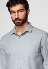 Homme aux cheveux noirs bouclés et à la barbe, portant une chemise gris clair à boutons avec une poche sur la poitrine, sur un fond uni.