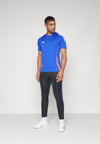 Blaues Sportshirt mit kurzen Ärmeln und orangen Akzenten. Schwarze, enganliegende Hose mit blauen Seitenstreifen. Weiße Sportschuhe.