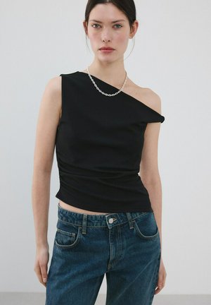 Žena v čiernom jednoramennom topu, so strieborným reťazovým náhrdelníkom a modrými džínsami s vysokým pásom, na jednoduchom bielom pozadí.