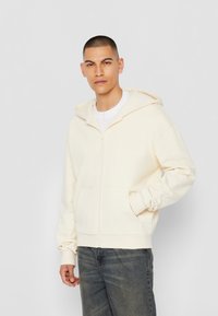 Haut à zip crème avec des poignets et une taille élastiques, doté d'une grande poche avant et d'une capuche doublée. Porté par-dessus un t-shirt blanc.