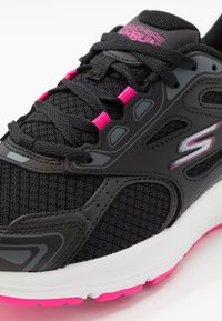 Czarne buty sportowe z siateczkowej tkaniny, różowe akcenty na sznurówkach i podeszwie oraz smukłe detale wzornicze wzdłuż boków. Biała śródpodeszwa.