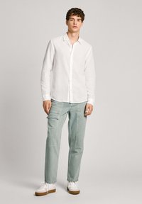 Camisa blanca de lino con botones combinada con pantalones cargo verde claro, que cuentan con múltiples bolsillos, y zapatillas blancas con suelas de goma marrón.