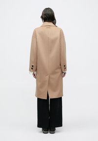 Barna gyapjúkabát laza fazonnal, ejtett vállakkal, nagy gombokkal és hátul sliccel, fekete bőszárú nadrággal párosítva.