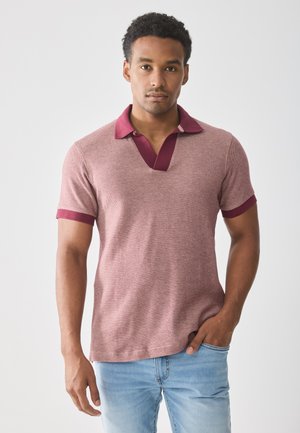 Man with short curly hair wearing a maroon-collared, short-sleeve knit shirt and light blue jeans, standing against a plain white background.