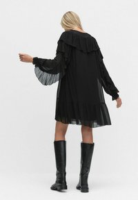 Kobieta w luźnej czarnej sukience z falbanami przy rękawach i u dołu, w połączeniu z czarnymi skórzanymi botkami do kolan, stojąca tyłem z lekko uniesionymi rękami.