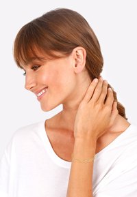 Gold chain bracelet worn on wrist, featuring oval links with textured accents. Model has brown hair, wearing a white t-shirt.