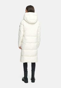Hvid pufferjakke med hætte, med et langt design, quiltet tekstur og sidelommer, båret over sorte leggings og støvler.