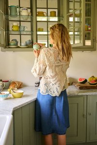 Woman in a blue skirt and cream lace top takes a green cup from a cabinet filled with colorful mugs and pitchers in a kitchen.