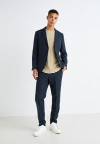 Completo blu navy realizzato in tessuto testurizzato, con giacca a un petto, pantaloni coordinati e sneakers bianche abbinate a una t-shirt beige.