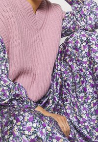 Růžový pletený svetr s výstřihem do V. Pod ním květinové šaty s fialovými, bílými a žlutými květinami na tmavém pozadí.