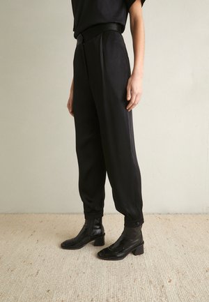 Personne portant un pantalon noir taille haute à coupe fuselée avec des poignets à boutons et des bottines noires à talons, debout sur un sol clair texturé.