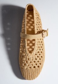 Chaussure plate tissée beige avec un bout rond et une bride à boucle sur le dessus, projetant des ombres à motifs sur une surface blanche.